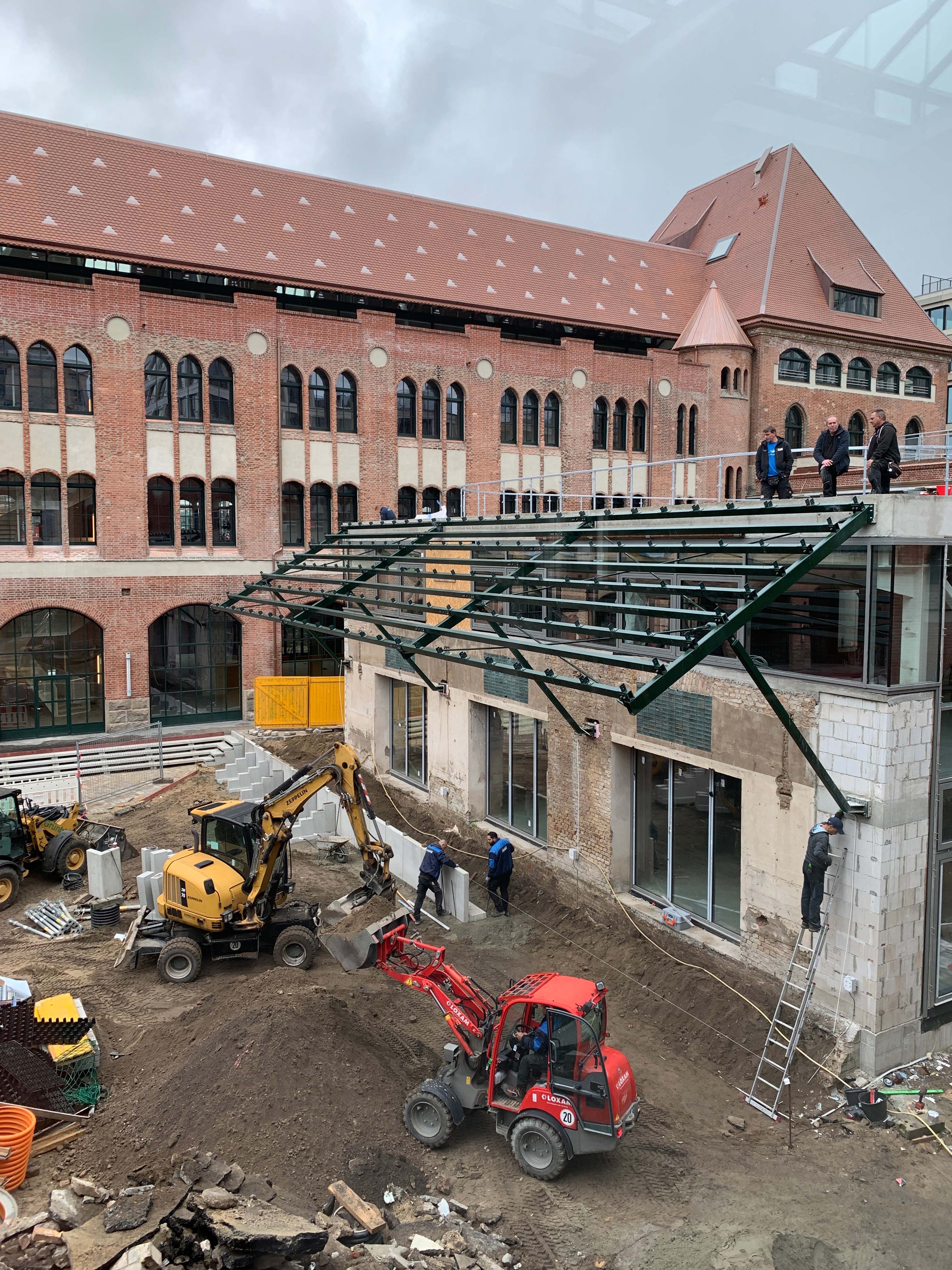 postbahnhof-offices-berlin-baufortschritt-blick-auf-beutel-APK Baustelle vor Backsteingebäude mit Bauarbeitern und Baggern, Blick auf die entstehende Struktur der Packkammer.
