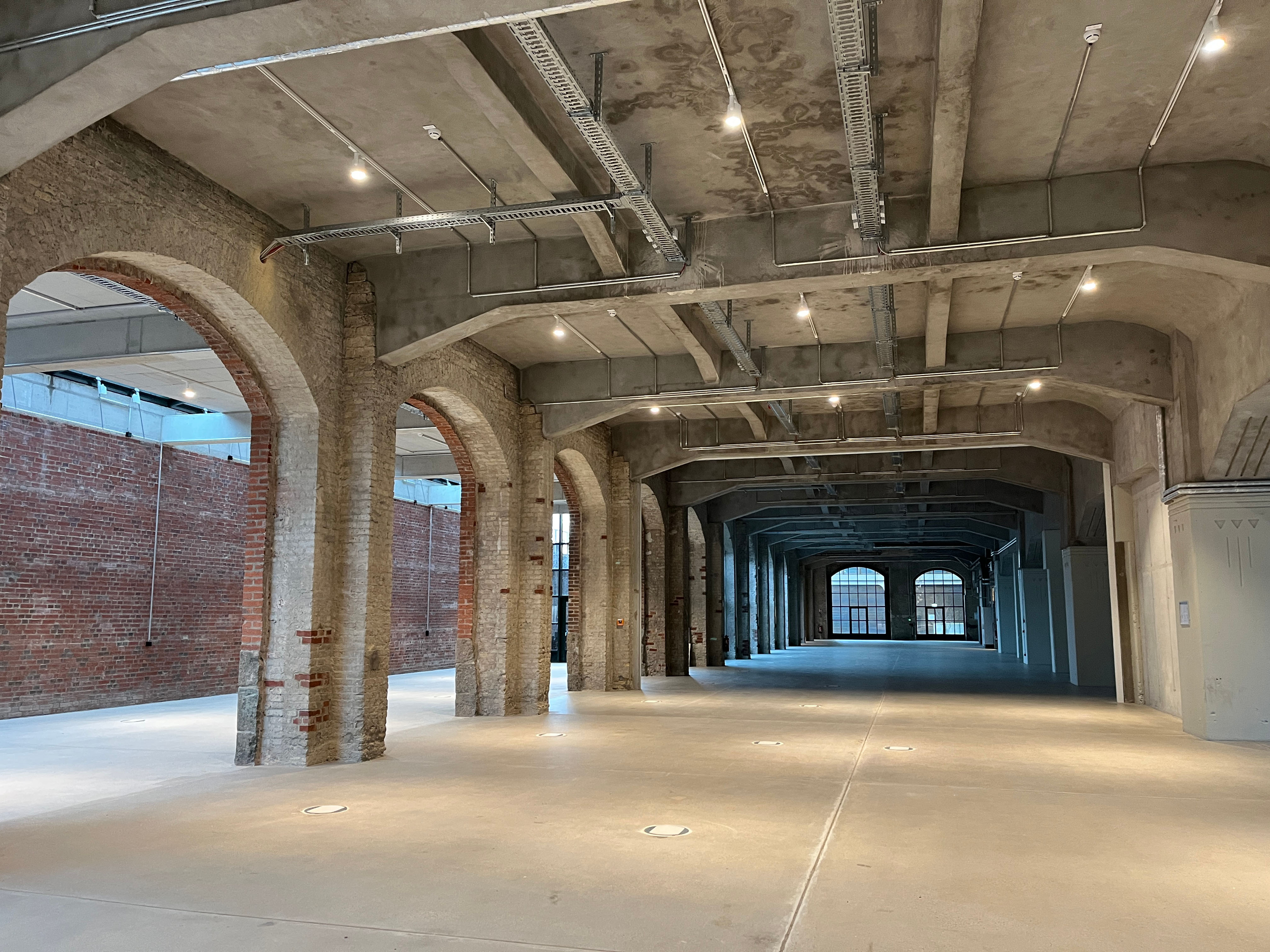 The construction progress at Postbahnhof Berlin shows the Abgangspackkammer on the ground floor with a large, open interior, high ceilings, evenly spaced columns, and natural daylight.