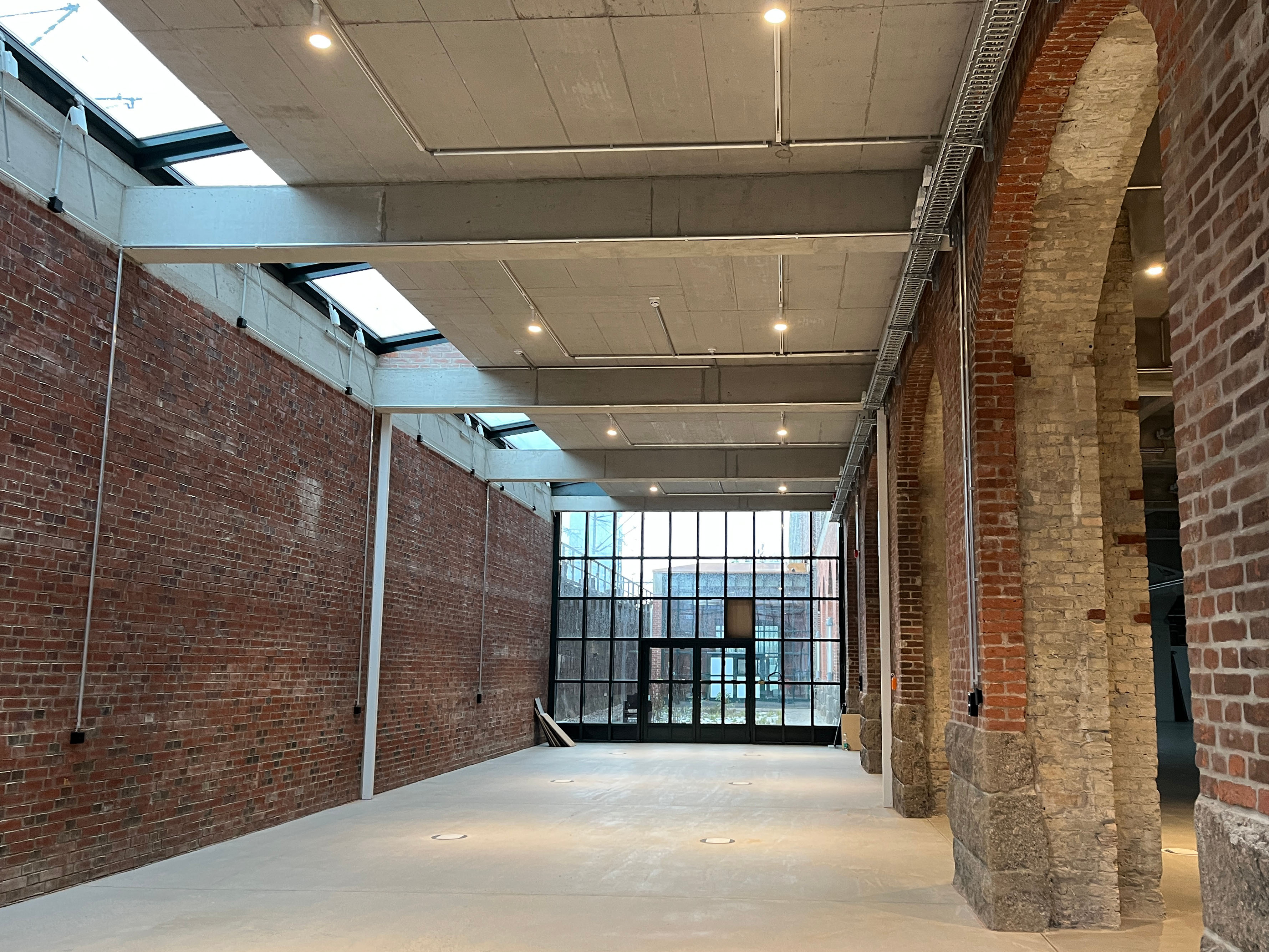 The construction progress at Postbahnhof Berlin shows the Abgangspackkammer on the ground floor with a long, empty corridor, exposed brick walls, structural columns, and a window front at the end of the space.