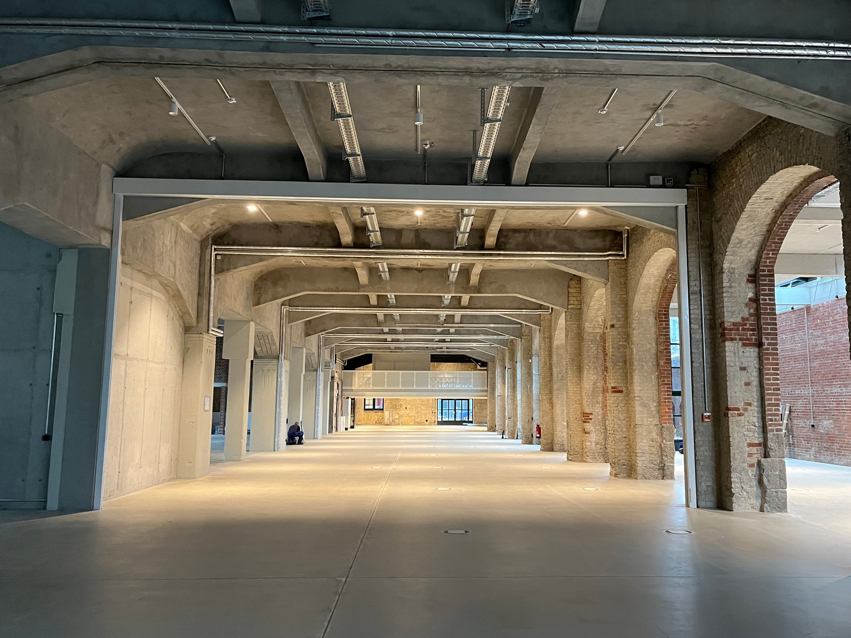 Der Baufortschritt im Postbahnhof Berlin zeigt die Abgangspackkammer im Erdgeschoss mit einem tiefen, leeren Innenraum, wiederkehrenden Pfeilern, hellem Boden und industrieller Deckenstruktur.