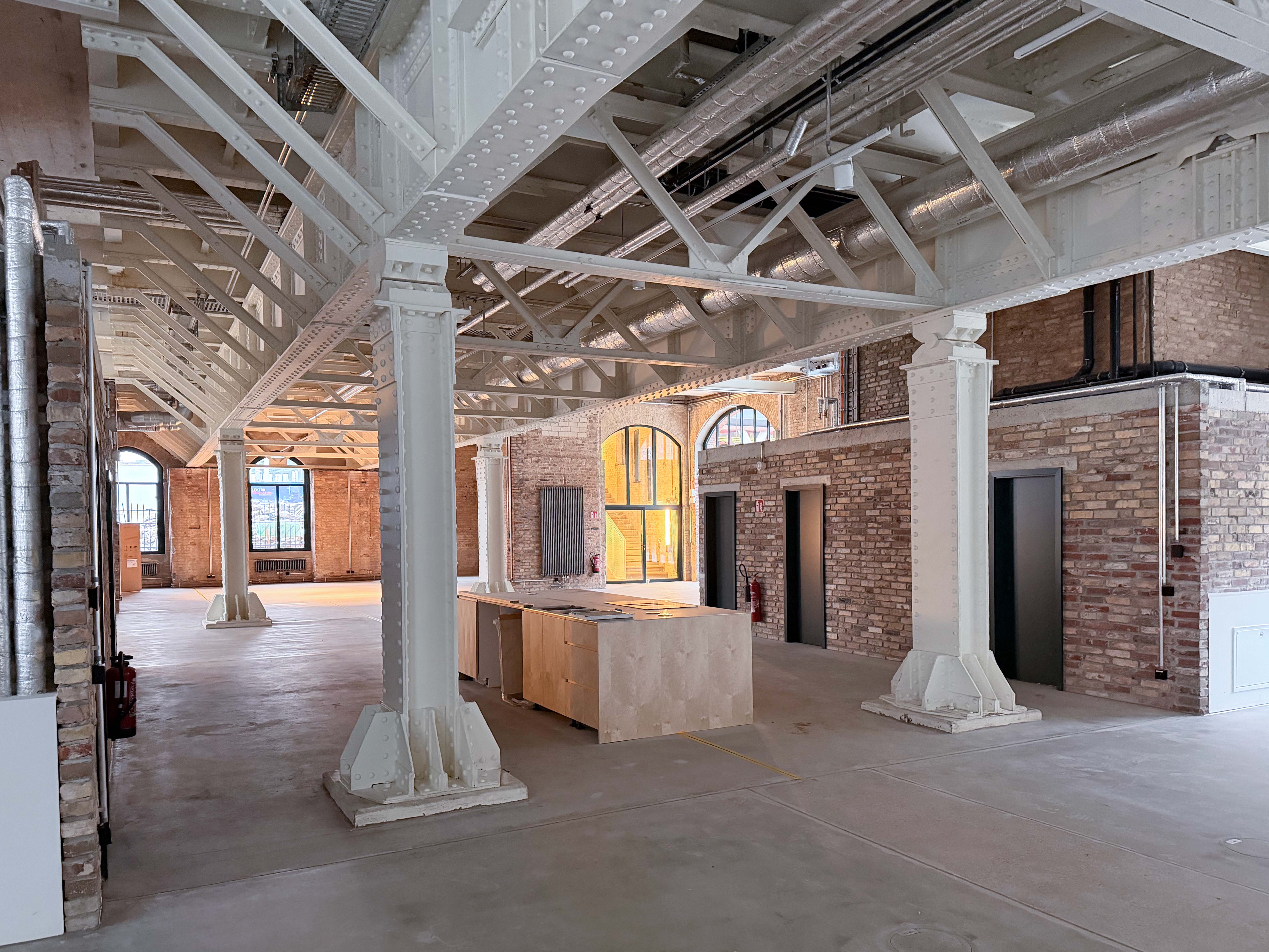 Der Baufortschritt im Postbahnhof Berlin zeigt die Eingangspackkammer im Erdgeschoss mit industriellen Stahlstützen, freigelegter Tragstruktur und neu entstehenden Einbauten.