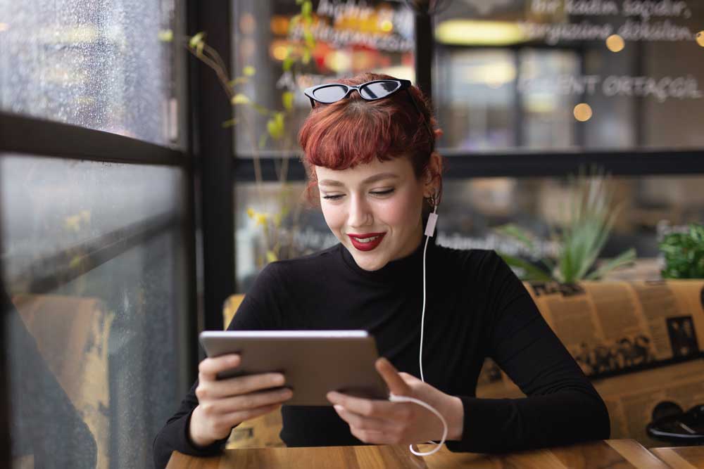 Eine Frau mit rotem Haar und Kopfhörern, die in einem Café ein Tablet benutzt.